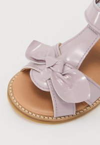 Sandale en cuir verni lavande avec un grand nœud décoratif, dotée d'une semelle intérieure en cuir beige et de bords surpiqués pour un détail supplémentaire.