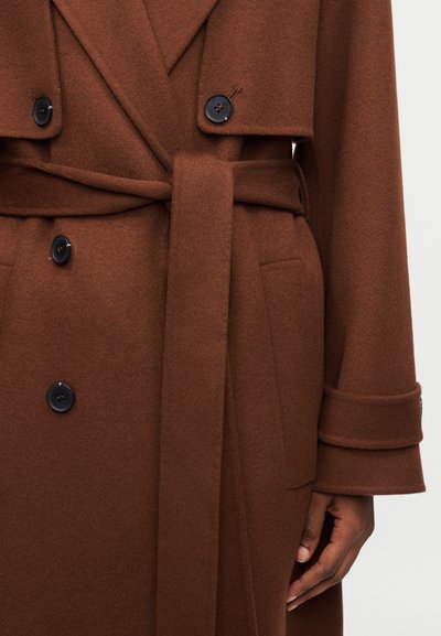 Καφέ μάλλινο παλτό διπλού στήθους με μαύρα κουμπιά, υφασμάτινη ζώνη δεμένη στη μέση, ιμάντες στις μανσέτες και το δεξί χέρι ακουμπισμένο δίπλα στην τσέπη.