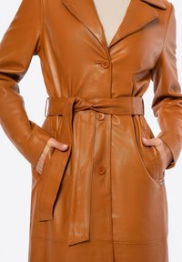 Hnědý kožený trench coat s páskem v pase, dvěma bočními kapsami a zapínáním na knoflíky. Obsahuje klopený límec a hladkou texturu.