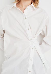 Weißes Button-Up-Hemd aus leichtem Baumwollstoff, mit klassischem Kragen, Vorderknöpfen und gerafftem Stoffdetail in der Taille.