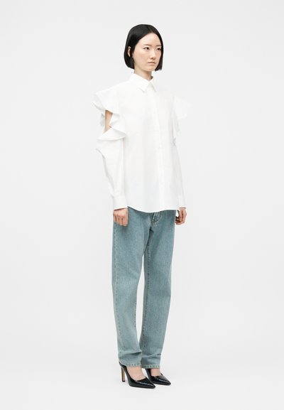 Femeie în picioare, purtând o bluză albă cu volane și umeri descoperiți, blugi albăstrui deschis și pantofi negri cu toc ascuțit, pe un fundal alb simplu.