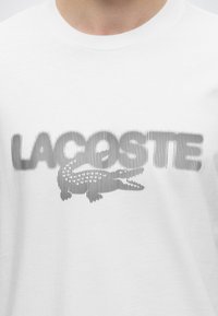 Λευκό T-shirt με ριγέ λογότυπο Lacoste και ένα στιλιζαρισμένο κροκόδειλο σε μαύρο εκτυπωμένο στο στήθος.