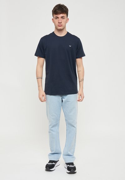 T-shirt en coton bleu marine à manches courtes et col rond, arborant un petit logo sur la poitrine, associé à un jean bleu clair et des baskets noires.