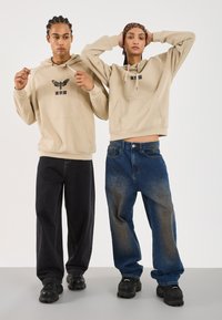 Beige hættetrøjer med trykte grafikker, parret med løse sorte og blå jeans. Begge bærer sorte sko med runde såler og teksturerede designs.