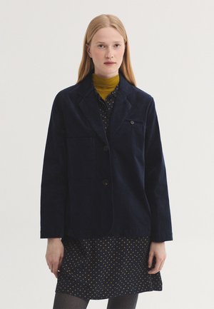 Jonge vrouw met steil blond haar draagt een donkerblauw blazer over een donkere jurk met gele stippen en een mosterdgele coltrui.