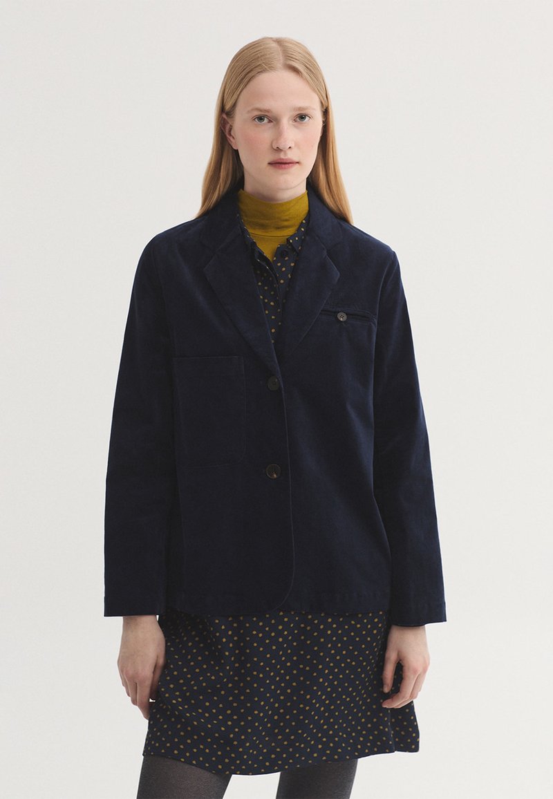 Jeune femme aux cheveux blonds et raides porte un blazer bleu foncé par-dessus une robe sombre à pois jaunes et un col roulé jaune moutarde.