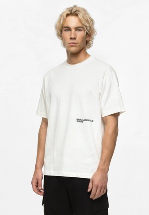 Jeune homme aux cheveux blonds portant un t-shirt blanc Karl Lagerfeld Jeans et un pantalon noir, debout devant un fond blanc uni.