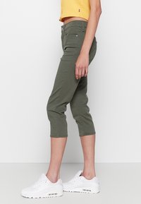 Oliwkowe rurki o kroju cropped z dopasowanym fasonem, z przednimi kieszeniami i gładką teksturą, zestawione z białymi sneakerami.