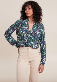 Bunte, gemusterte Bluse mit langen Ärmeln und Kragen, kombiniert mit hellbeigen Hosen mit Knopfakzenten.