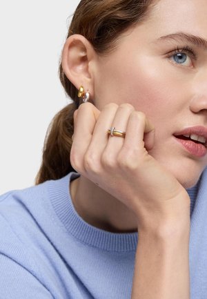 Giovane donna che indossa orecchini a cerchio in oro e argento e un anello in oro e argento, con il mento appoggiato sulla mano, indossa un maglione blu visibile.