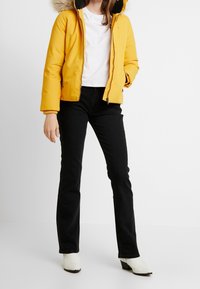 Geel pufferjack met een bontvoering bij de capuchon, een ritssluiting aan de voorkant en ribgebreide manchetten, gecombineerd met zwarte jeans en witte haklaarzen.