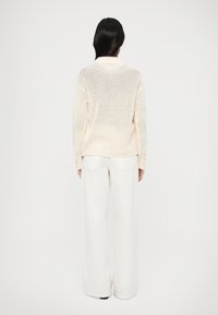 Πλεκτό πουλόβερ σε ανοιχτό κρεμ με υφασμένο σχέδιο, συνδυασμένο με λευκό παντελόνι φαρδιάς γραμμής. Το πουλόβερ έχει γιακά και ριμπ μανσέτες.
