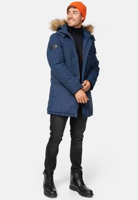 Manteau d'hiver bleu marine avec une capuche doublée en fourrure, fermeture éclair à l'avant et grandes poches, associé à un pantalon noir et des bottes noires à lacets. Bonnet orange.