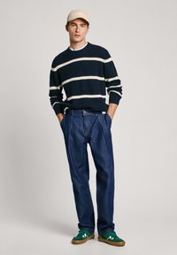 Maglione a righe navy con righe crema, pantaloni in denim blu e sneakers verdi. Vestibilità non strutturata; berretto con logo del brand, sfondo chiaro.