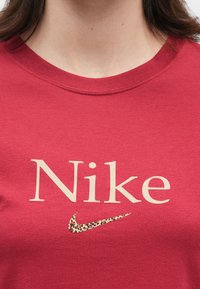 Κοντινό πλάνο ατόμου που φοράει κόκκινο πουκάμισο με μπεζ κείμενο "Nike" και λογότυπο σφύρας Nike με λεοπάρ σχέδιο στο στήθος.