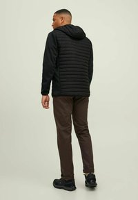 Schwarze isolierte Kapuzenjacke mit gestepptem Frontbereich und glatten Ärmeln, kombiniert mit braunen Hosen und schwarzen Sneakern, von hinten betrachtet.