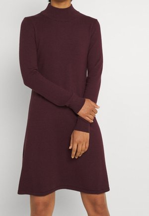 Person wearing a long-sleeve, knee-length, maroon mock neck dress with hands gently clasped in front against a plain background.