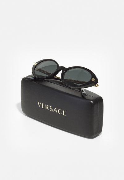 Svarta ovala solglasögon med mörka linser, vilande på en svart hård förpackning prydd med "VERSACE" i guldbokstäver. Guldaccenter på ramen.