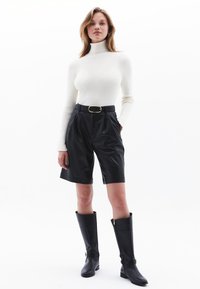 Schwarze plissierte Shorts mit Taschen, kombiniert mit einem gerippten weißen Rollkragenpullover. Das Modell trägt kniehohe schwarze Stiefel mit goldenen Reißverschluss- und Schnallenakzenten.