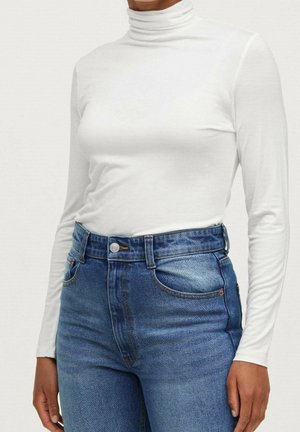 Osoba ubrana w dopasowany biały golf i niebieskie dżinsy z wysokim stanem, stojąca z ramionami swobodnie opuszczonymi wzdłuż ciała.