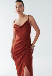 Rostfärgad klänning med polokrage i slät tyg med sidodrapering och slits högt upp på låret. Tunna axelband och minimalistiska guldsmycken.