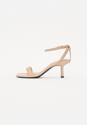 Beige åben tå højhælet sandal med tynd ankelrem og guldlås på hvid baggrund.
