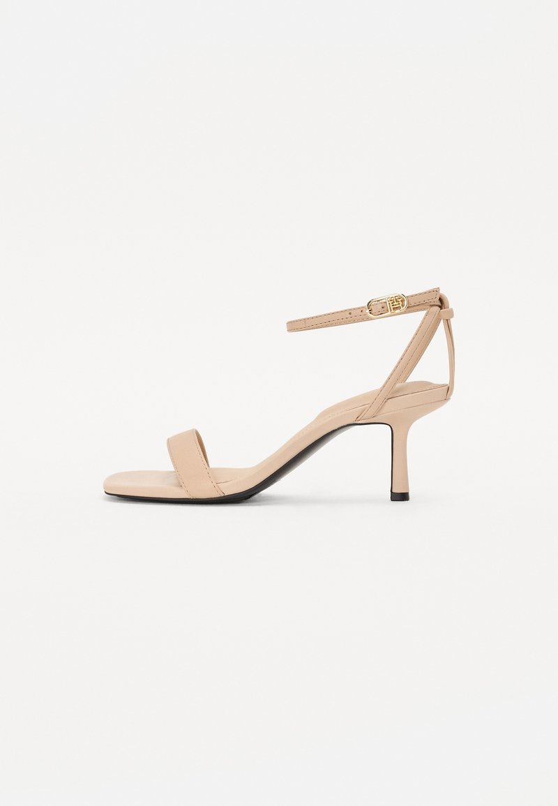 Sandale beige à talon haut ouvert à l'avant avec fine bride à la cheville et boucle dorée sur fond blanc.