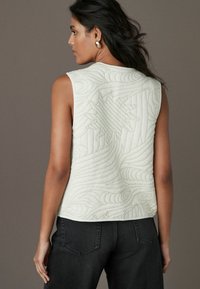 Ærmeløs quiltet top i lys creme med teksturerede svulmønstre på ryggen og en rund halsudskæring. Sammen med mørke jeans.