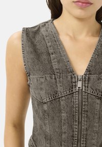 Chaleco de mezclilla en gris con diseño sin mangas, escote en V profundo, cierre delantero con cremallera y costuras estructuradas. Textura suave con un acabado desgastado.