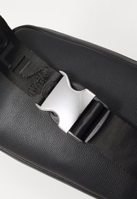 Schwarze Ledertasche mit einer strukturierten Oberfläche, ausgestattet mit einer metallischen silbernen Schnalle und einem geflochtenen schwarzen Tragegurt, der auffällige Schriftzüge zeigt.