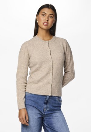 Beige strikket cardigan med rund hals og frontknapp-lukking. Har lange ermer og en myk tekstur, kombinert med denimjeans.