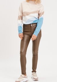 Strickpullover mit beigen, braunen und blauen Wellenmustern; enganliegende braune Hose; hellfarbene Sneakers mit dicker Sohle.