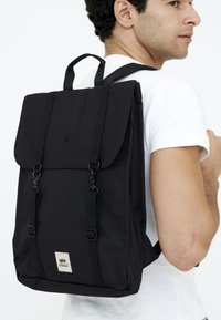 Svart ryggsäck i hållbart tyg, med klaffstängning, justerbara remmar och en liten logopatch på framsidan.