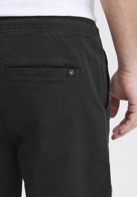 Le pantalon en coton noir dispose d'une poche arrière avec une ouverture horizontale et un accent de logo. La taille est élastique pour plus de confort et de flexibilité.
