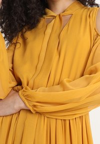 Sukienka w musztardowym kolorze wykonana z lekkiego, teksturowanego materiału. Posiada długie, bufiaste rękawy oraz dekolt w kształcie łezki z detalem w postaci wiązania.