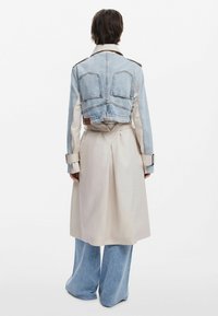 Długi beżowy trencz na lekko niebieską kurtkę dżinsową z krótkim rękawem. Zawiera duże tylne kieszenie oraz szerokie niebieskie dżinsowe spodnie.