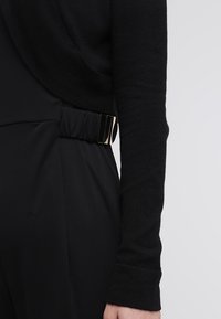 Gros plan d'une personne portant un pantalon noir avec une taille élastique et une boucle dorée, ainsi qu'un haut noir à manches longues sur un fond clair.