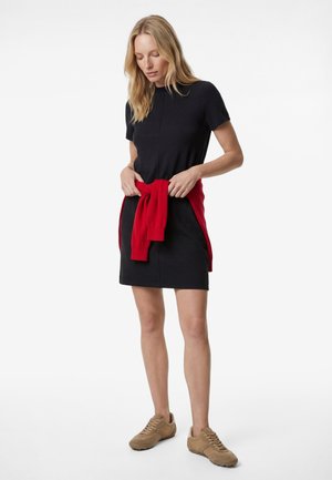 Blonde Frau trägt ein schwarzes kurzärmeliges Kleid mit einem roten Pullover, der um ihre Taille gebunden ist, und beige Sneaker, steht vor einem weißen Hintergrund.