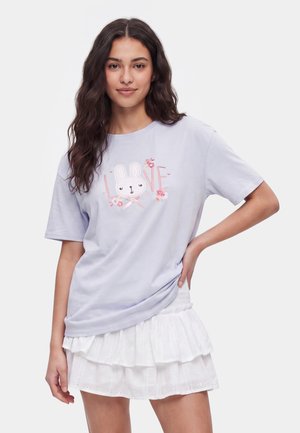 Kvinde med langt mørkt hår iført en lavendelfarvet t-shirt med en kanin og teksten "LOVE", kombineret med en hvid lagdelt mini nederdel med hulmønster.