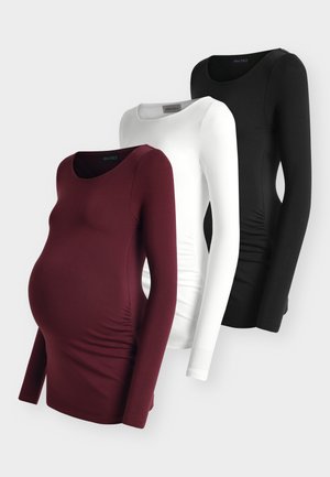 Donna incinta che indossa una maglia bianca a maniche lunghe, jeans scuri, tacchi aperti neri e porta una borsa a tracolla nera su uno sfondo neutro.