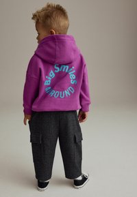 Lila Kapuzenpullover mit blauen Grafiken "Big Smiles AIRound" auf der Rückseite, kombiniert mit schwarz-karierter Cargo-Hose und schwarzen Sneakern.