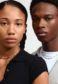 Två personer står nära varandra. Den ena har på sig en svart ribbad topp; den andra bär en ljusgrå zip-up och en vit skjorta. De har varsin pärlband runt halsen.