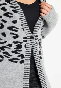 Grå cardigan med svart leopardtryck och randiga accentfärger, med en knappstängning och ribbade ärmslut, uppvisad över en svart topp.