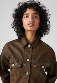 Brun denimjakke med krage, sølvknapper, brystlommer og en rød merkelapp. Strukturert finish med en figursydd silhuett.