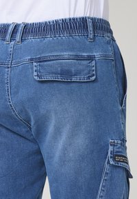 Modre denim hlače s cargo krojem, ki vsebujejo raztegljiv pas, zadnji žep in stranski cargo žep z črno oznako. Gladka tekstura.