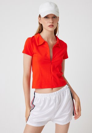 Korte rode polo met rits en kraag, gecombineerd met witte shorts met zwarte en rode zijkanten, alles tegen een neutrale achtergrond.