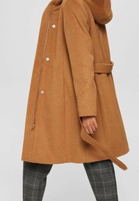 Vrouw in een camelkleurige wollen jas met knopen, rits en riem over grijs geruite broek tegen een effen achtergrond.