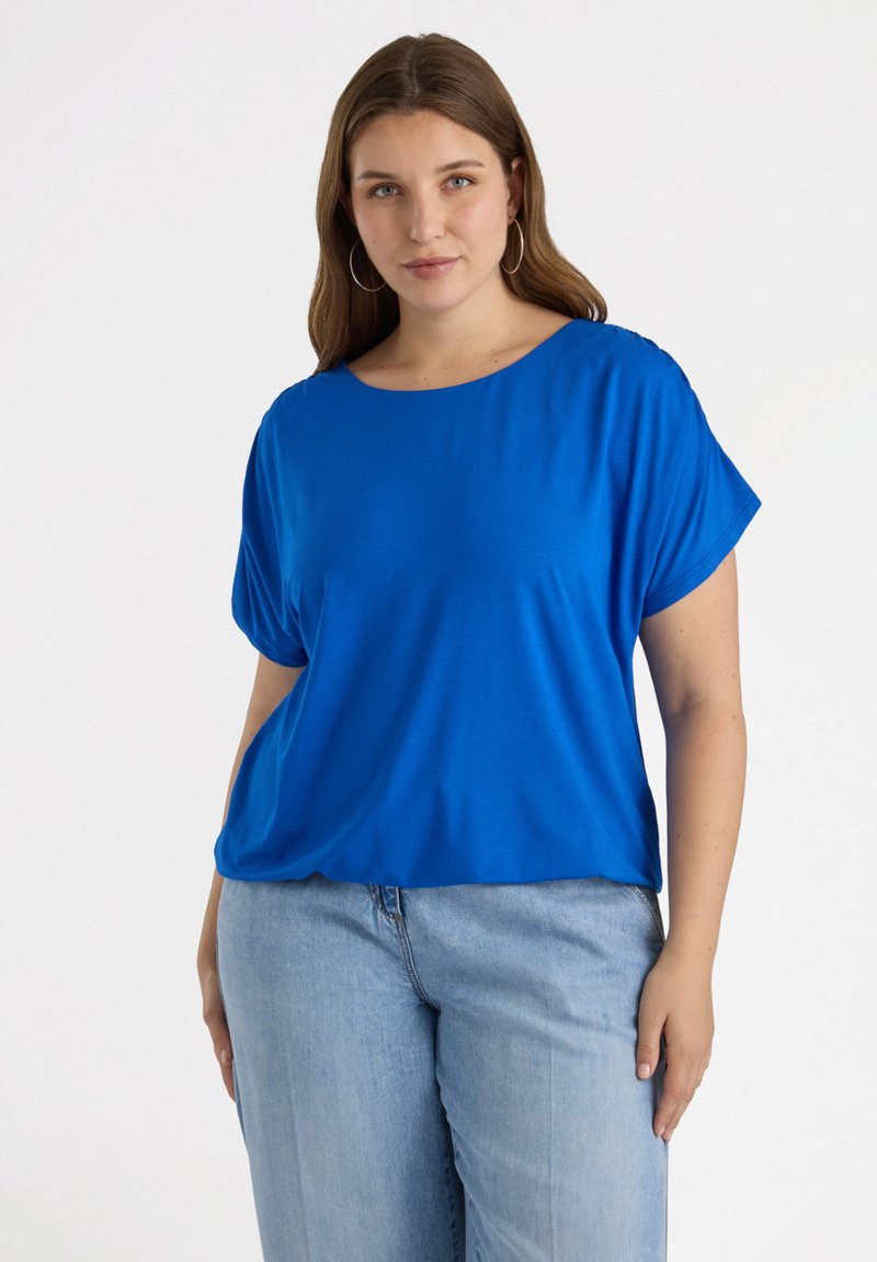 Blaues kurzärmliges Oberteil mit lockerer Passform und weitem Ausschnitt, aus glattem Stoff gefertigt. Kombiniert mit hellblauen Jeans, die einen lässigen Look unterstreichen.