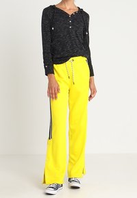 Person som bär ljusgula vida byxor med sidostråk och svart-vita sneakers, tillsammans med en svart huvtröja med lång ärm.