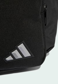 Mochila de tela negra con superficie texturizada y un logo triangular reflectante en gris. Compartimento con cremallera visible en el lateral.